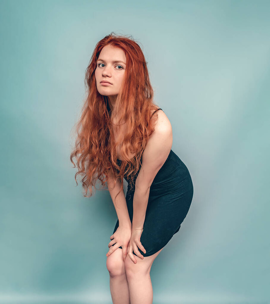 Tired redhead woman posing in studio on gray background. Long hair, black dress - Photo, Image
