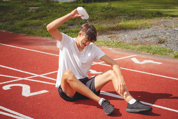 Jovem atleta refresca-se com água depois de um duro treino sobre o atlético oval no calor intenso. Treino de resistência. Adolescente de cabelos castanhos. - Foto, Imagem