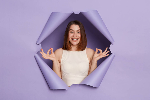 Smiling calm brunette woman breaking through purple paper hole wearing casual white top feeling harmony practicing yoga keeps hands in mudra gesture. - Photo, Image