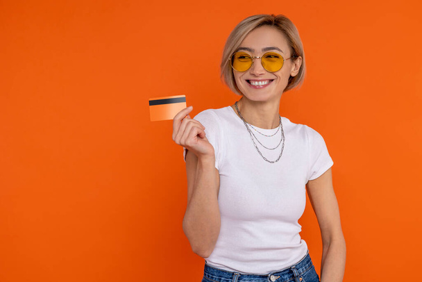 Charming smiling happy woman in white t shirt showing credit card banking isolated over orange background. - Photo, image