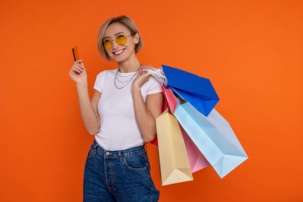 Satisfied woman in white t shirt and jeans holding shopping bags and credit card isolated over orange background. - 写真・画像
