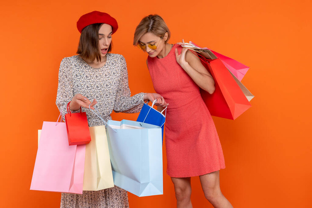 Erwachsene Freundinnen schauen in Einkaufstüten vor orangefarbenem Hintergrund. - Foto, Bild