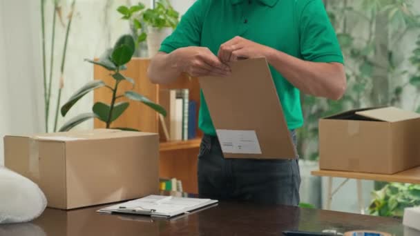 Υπάλληλος ταχυδρομείου με πράσινο ενιαίο έγγραφο συσκευασίας σε φάκελο και τη διεύθυνση γραφής σε αυτό πριν από την αποστολή στον πελάτη στο γραφείο - Πλάνα, βίντεο