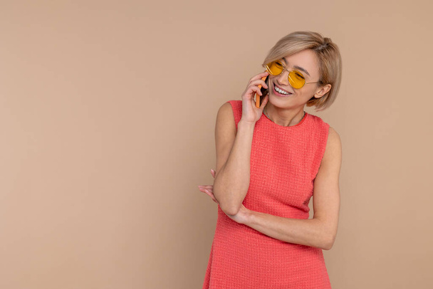 Happy smiling woman in dress and sunglasses talking mobile phone isolated over beige background. - Fotó, kép