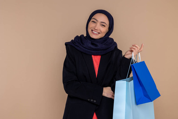 Smiling Muslim woman in hijab standing with shopping bags isolated over beige background. - Foto, immagini