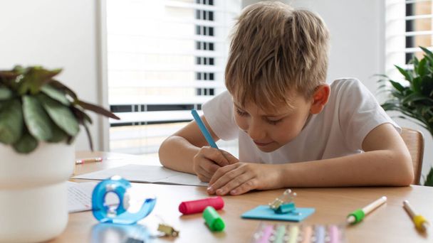 A cute boy does his homework at the table at home, writes in a notebook. Home schooling, elementary school. The concept of childrens education - Foto, Bild