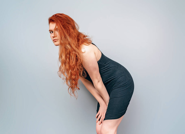 Visão lateral da menina com cabelos longos vermelhos inclinados para a frente. Vestido curto preto. Estúdio foto - Foto, Imagem