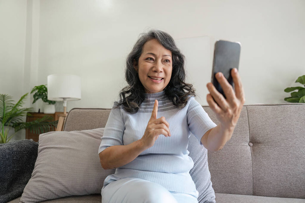 Happy mature middle aged woman waving hand holding mobile phone video conference calling by social distance virtual family online chat meeting sitting on couch at home. - Photo, image