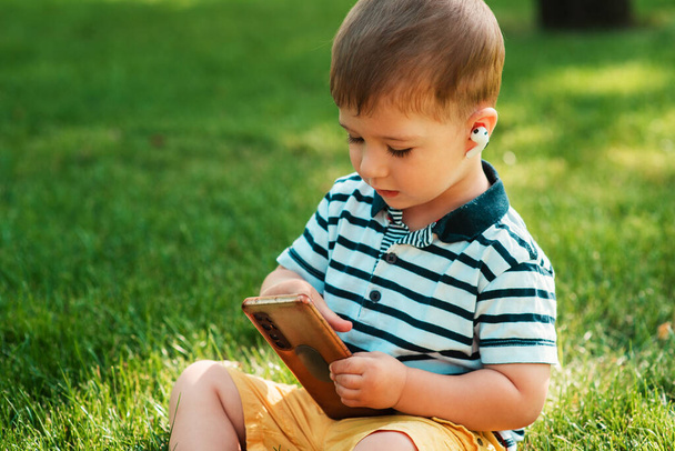 A cute European-looking boy studies the workings of a smartphone with wireless headphones in his ears on the green grass. A young child uses gadgets. Entertainment and leisure time of a child - Fotografie, Obrázek