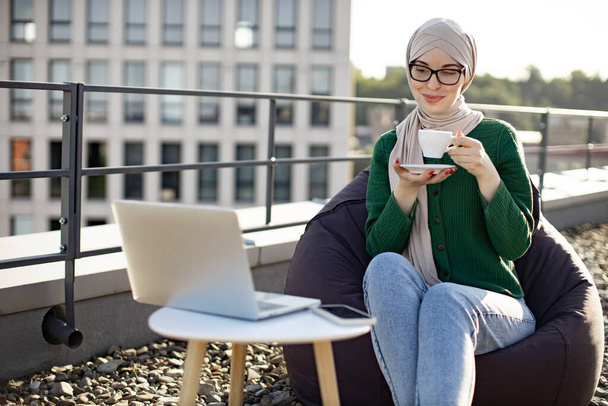 Cheerful woman in headscarf having cup of hot drink while resting in beanbag chair near coffee table with gadgets. Young muslim adult increasing creativity while staying on panoramic terrace at noon. - Фото, зображення
