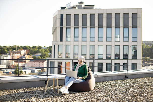 Wide view of attractive muslim woman in casual clothes and hijab working on laptop while sitting in beanbag. Thoughtful young professional reading report via digital device on panoramic terrace. - Foto, Imagem