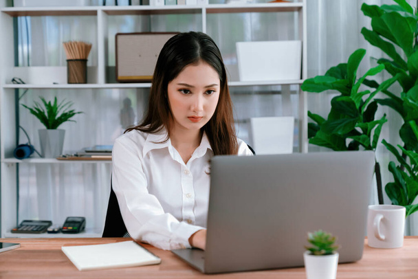 Молодая азиатская восторженная деловая женщина за современным офисным столом использует ноутбук для работы с чашкой кофе. Усердная и привлекательная офисная леди, работающая над ноутбуком в офисе. - Фото, изображение