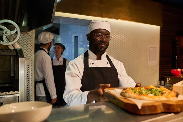 Young serious African American male chef in uniform putting wooden board or tray with appetizing Italian pizza on counter against colleagues - 写真・画像