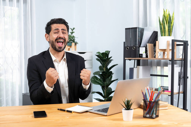 Siyah takım elbise giymiş heyecanlı ve mutlu bir iş adamı başarılı bir iş tanıtımı, enerji ve neşe kazandıktan sonra ofis masasında kutlama jesti olarak elini kaldırır. Coşkulu - Fotoğraf, Görsel