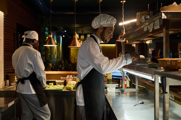 Zijaanzicht van jonge mannelijke chef-kok in uniform die schotel klaarmaakt om te serveren terwijl hij achter de toonbank staat tegen zijn collega die verse groenten hakt - Foto, afbeelding