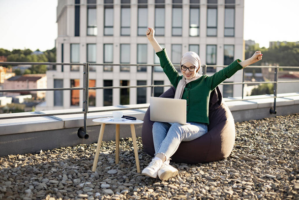 Emotional female in hijab raising arms in victory while looking at computer screen on rooftop of building. Cheerful muslim woman in spectacles celebrating successful purchase via internet outdoors. - Photo, Image