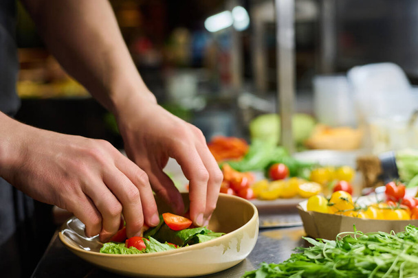 Genç erkek aşçının ya da şefin elleri masada servis yapmaya hazırlanırken taze sebze salatası malzemelerini tabağa koyuyor. - Fotoğraf, Görsel