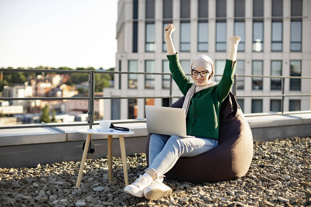 Emotional female in hijab raising arms in victory while looking at computer screen on rooftop of building. Cheerful muslim woman in spectacles celebrating successful purchase via internet outdoors. - Foto, Imagen