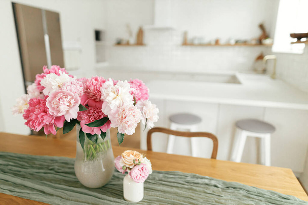 Modern kitchen interior. Beautiful peonies in vase on wooden table on background of stylish white kitchen with appliances in new scandinavian house.Summer floral arrangement - Фото, изображение