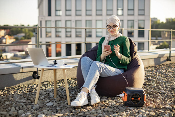 Focused arabian lady in hijab holding cell phone connected to portable power station via charging cord on roof terrace. Efficient freelancer working remotely using eco-friendly energy source outside. - Foto, Imagem