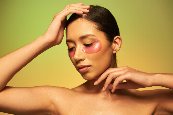 Schönheitskampagne, junge Asiatin mit brünetten Haaren und sauberer Haut posiert mit gesichtsnahen Händen auf grünem Hintergrund, nackten Schultern, feuchtigkeitsspendenden Augenflecken, glühender Haut  - Foto, Bild