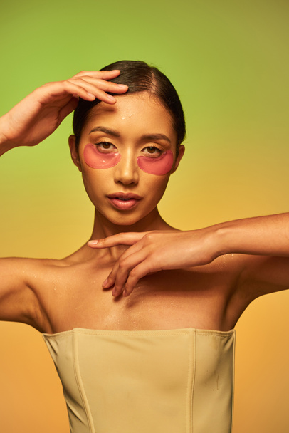 Hautpflege-Kampagne, junge asiatische Frau mit brünetten Haaren und sauberer Haut posiert und blickt in die Kamera auf grünem Hintergrund, nackte Schultern, feuchtigkeitsspendende Augenflecken, glühende Haut  - Foto, Bild