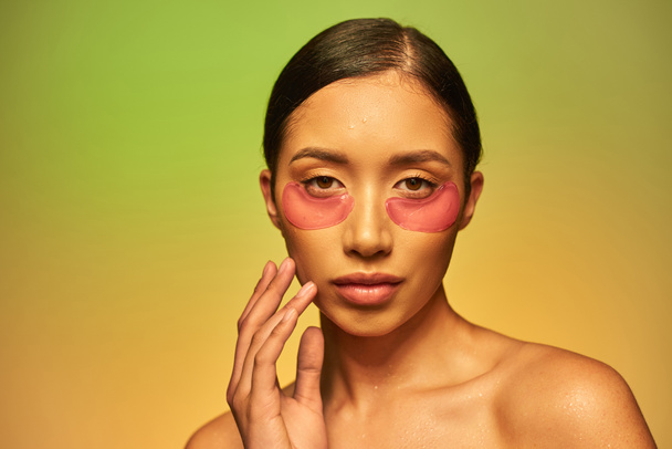 Cilt bakımı kampanyası, esmer saçlı ve temiz tenli genç Asyalı kadın yanağa dokunuyor ve yeşil arka planda kameraya bakıyor, çıplak omuzlar, nemlendirici göz bantları, parlayan cilt.  - Fotoğraf, Görsel