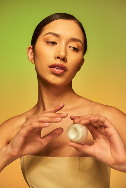 beauty campaign, young asian woman with bare shoulders holding cosmetic jar with face cream on green background, brunette hair, beauty industry, glowing skin, skin care concept  - Photo, Image