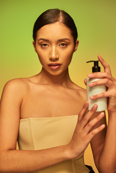 termék bemutatása, bőrápolási termék, fiatal ázsiai nő csupasz vállak gazdaság kozmetikai üveg testápoló és pózol zöld háttér, ragyogó bőr koncepció  - Fotó, kép