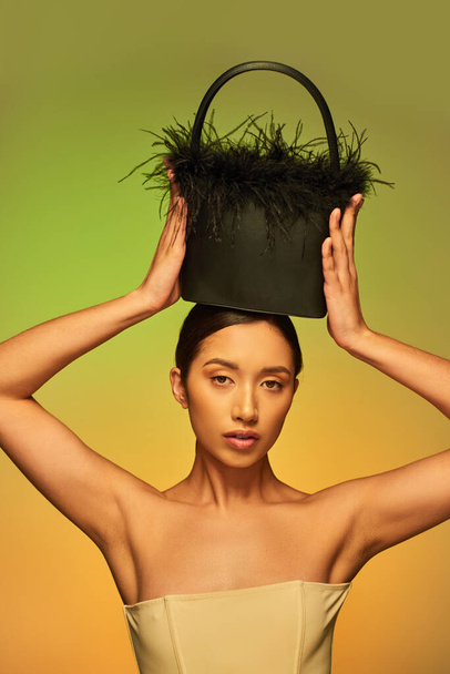 beauty and style, brunette asian woman with bare shoulders posing with feather purse on head on green background, gradient, fashion statement, glowing skin, natural beauty, young model  - Fotografie, Obrázek