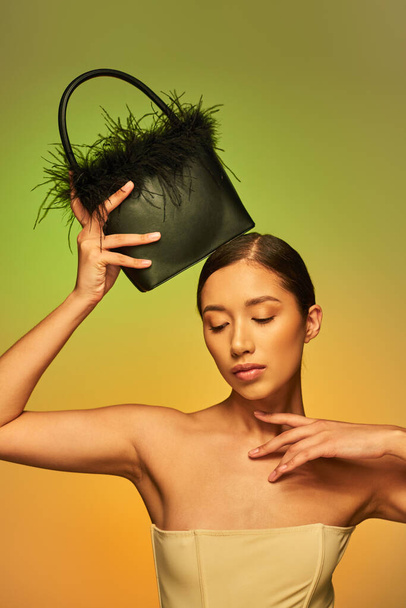 krása a styl, brunetka asijská žena s holými rameny pózující s péřovou kabelkou na zeleném pozadí, ruka v blízkosti obličeje, gradient, módní prohlášení, zářící kůže, přírodní krása, mladý model  - Fotografie, Obrázek