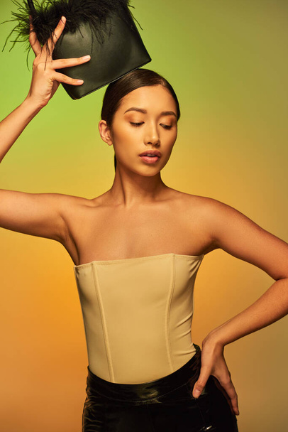beauty and style, brunette asian woman with bare shoulders posing with feather purse and hand on hip on green background, gradient, fashion statement, glowing skin, natural beauty, young model  - Φωτογραφία, εικόνα