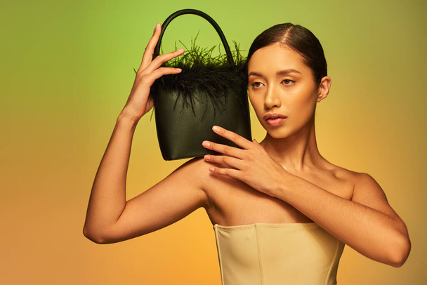 fashion choices, brunette asian woman with bare shoulders posing with feather purse and looking away on green background, gradient, fashion statement, glowing skin, natural beauty, young model  - Photo, Image