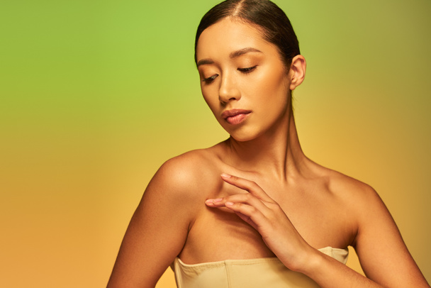 Hautpflege und Schönheit, asiatische Frau mit brünetten Haaren und nackten Schultern posiert auf Gradienten Hintergrund, grün und orange, Hautpflege, glühende Haut, natürliche Schönheit, junges Modell  - Foto, Bild