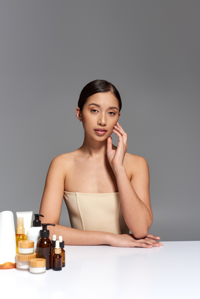 product presentation, skin care, young asian model with brunette hair posing near different beauty products on grey background, glowing and heathy skin, beauty campaign, facial treatment concept  - Foto, Imagem