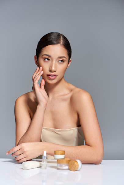Cilt bakımı sunumu, gri arka planda farklı güzellik ürünlerinin yanında çıplak omuzlu Asyalı genç bir kadın, parıldayan ve sağlıklı cilt, güzellik kampanyası, yüz bakımı konsepti  - Fotoğraf, Görsel