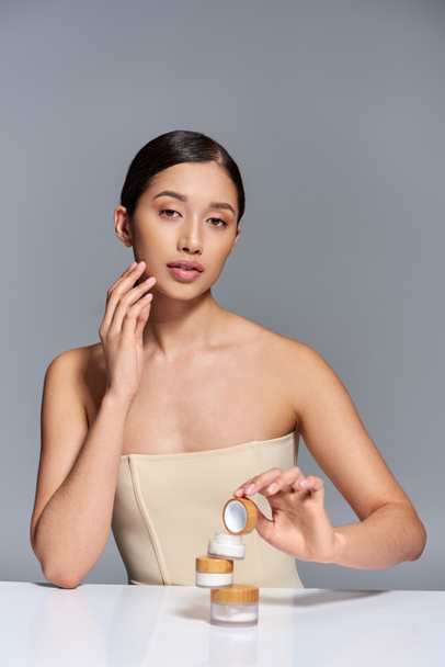 product presentation, skin care, young asian model with brunette hair holding face cream on grey background, glowing and heathy skin, beauty campaign, facial treatment, conceptual  - Photo, Image