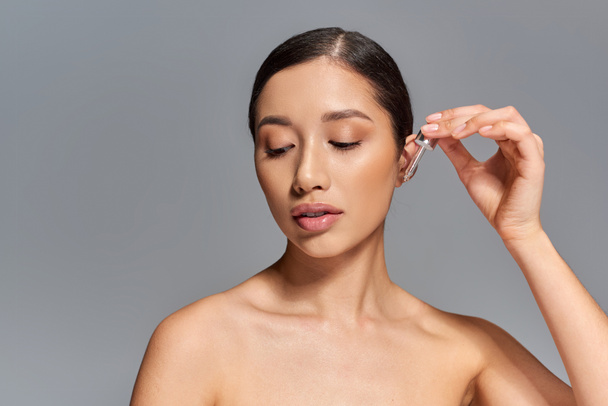 beauty campaign, skin care, young asian woman with brunette hair applying serum with pipette on grey background, glowing and heathy skin, facial treatment concept, looking away - Photo, Image