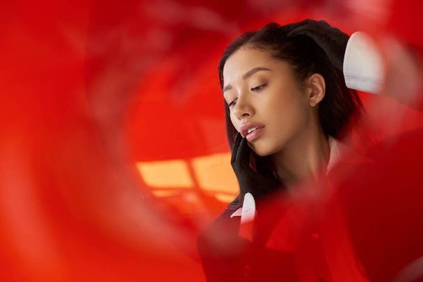 modern kadın, beyaz gömlekli ve ceketli genç Asyalı model kırmızı cam arkasında eldivenlerle poz veriyor, kişisel stil, gençlik eğilimi, kavramsal, dokunaklı boyun ve stüdyoda poz veriyor. - Fotoğraf, Görsel