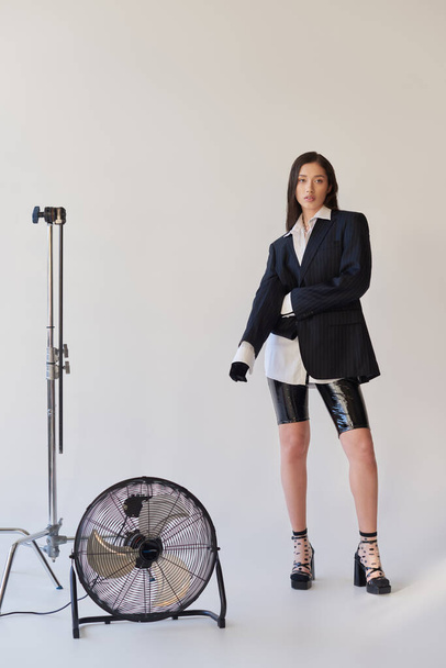 fashion forward, studio photography, young asian woman in stylish look posing on grey background, blazer, white shirt and latex shorts, standing near electric fan, personal style, full length  - Valokuva, kuva