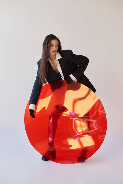 Mode und Stil, Studiofotografie, junges asiatisches Model in trendiger Kleidung posiert in der Nähe von rotem Rundglas, grauem Hintergrund, Blazer und Latex-Shorts, persönlicher Stil, volle Länge  - Foto, Bild