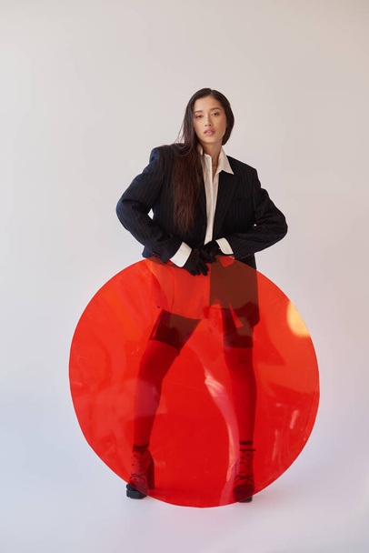 edgy style, studio photography, young asian woman in stylish look posing near red round shaped glass, grey background, blazer and latex shorts, youthful fashion, full length  - Photo, Image