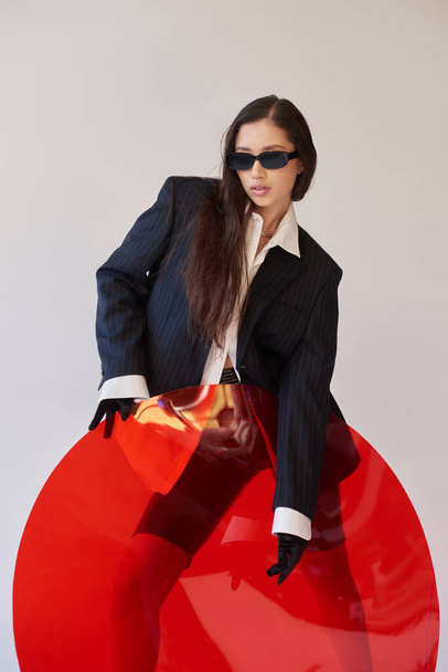 edgy style, studio photography, young asian woman in stylish look and sunglasses posing near red round shaped glass, grey background, blazer and latex shorts, youthful fashion, cool style  - Photo, Image
