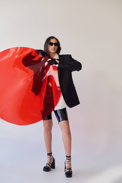 pretty asian model in stylish look and sunglasses posing holding red round shaped glass, grey background, blazer and latex shorts, youthful and modern woman, fashion statement, studio photography  - Foto, imagen