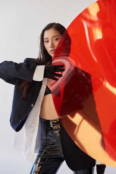 beautiful asian woman in trendy outfit holding red round shaped glass, grey background, blazer and latex shorts, youthful model, fashion forward, studio photography, conceptual  - Foto, Bild