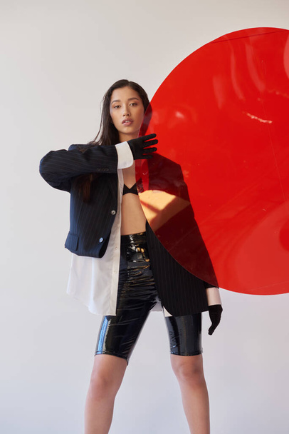 красива азіатська жінка в модному вбранні, що тримає червоне кругле скло, сірий фон, блейзер і чорні латексні шорти, молода модель в рукавичках, мода вперед, студійна фотографія, концептуальна
  - Фото, зображення