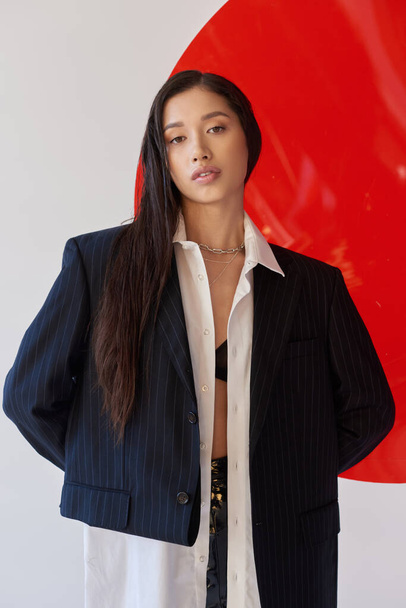 fashion forward, pretty asian woman in trendy outfit posing near red glass on grey background, blazer and black latex shorts, youthful model, studio photography, conceptual  - Φωτογραφία, εικόνα