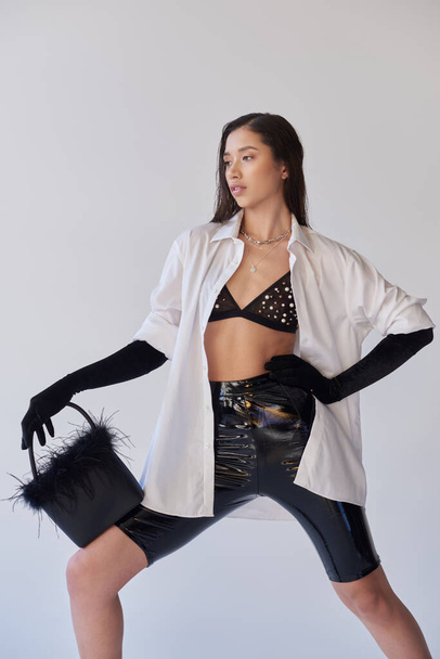 edgy style, fashion statement, asian woman posing with feathered handbag on grey background, young model looking away, black gloves and white shirt, latex style, conceptual, personal style  - Foto, immagini