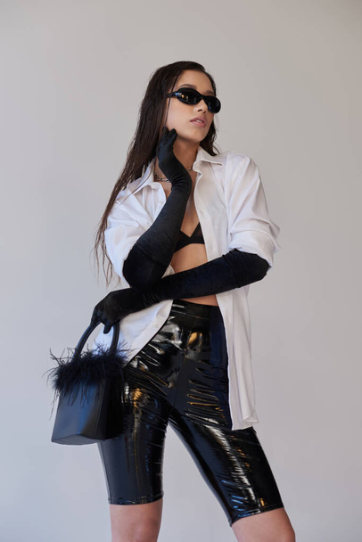bold style, fashion statement, asian woman in sunglasses posing with feathered handbag on grey background, young model in latex shorts, black gloves and white shirt, conceptual  - Foto, Imagem