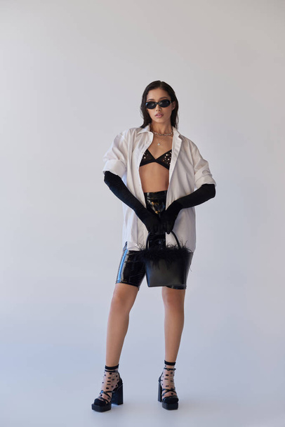 trendy look, fashion statement, brunette asian woman in sunglasses posing with feathered purse on grey background, model in latex shorts, black gloves and white shirt, youth, full length  - Fotografie, Obrázek